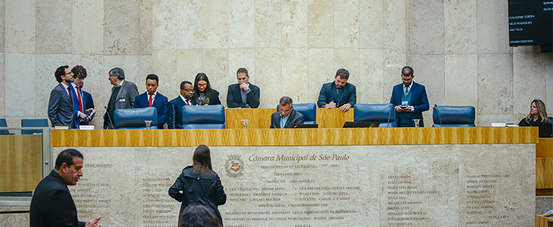 =Pessoas conversam e consultam celulares junto à mesa do Plenário da Câmara Municipal de São Paulo.