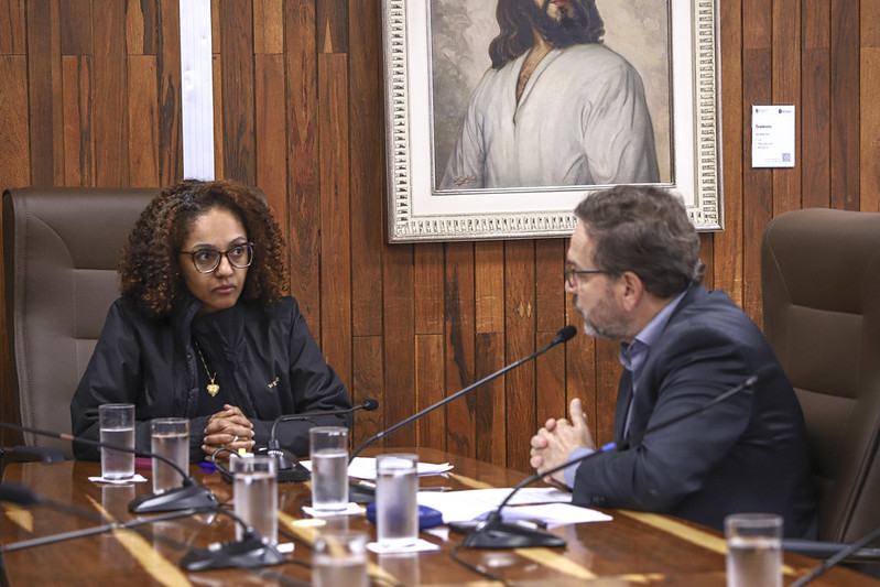 =Mulher e homem conversam diante de microfones em uma mesa de reunião.